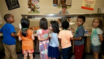 Children lined up in front of cafeteria screened buffet serving table.